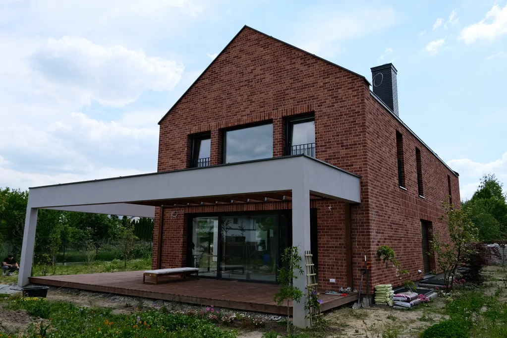 Widok od strony ogrodu na tylną elewację domu z ceglaną okładziną, zadaszonym tarasem i drewnianym podestem