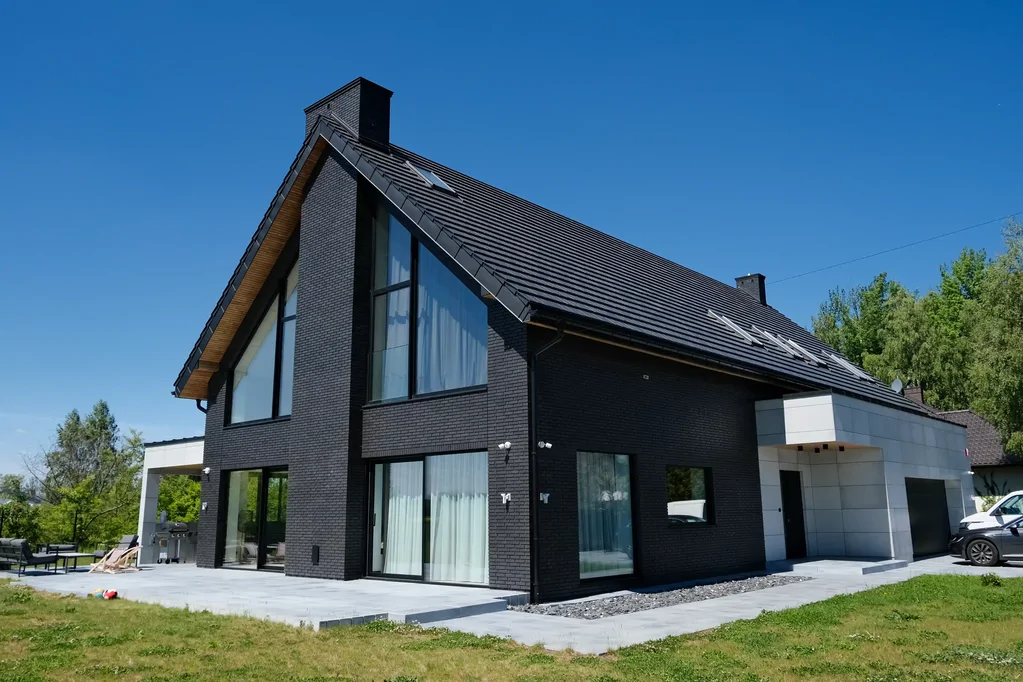 Widok z niskiego pułapu: nowoczesny dom z ciemnej cegły, przeszklenia szczytowe i bryła garażu z jasnych płyt.