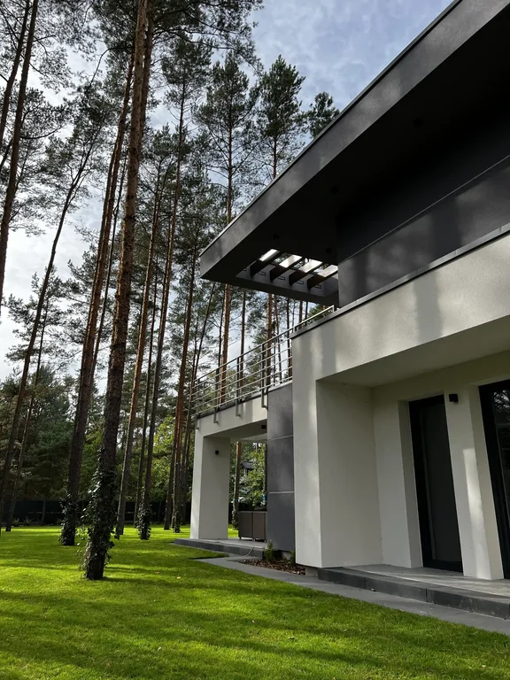 Detal elewacji bocznej: balkon ze stalową balustradą, duży okap i pergola, sosny w tle