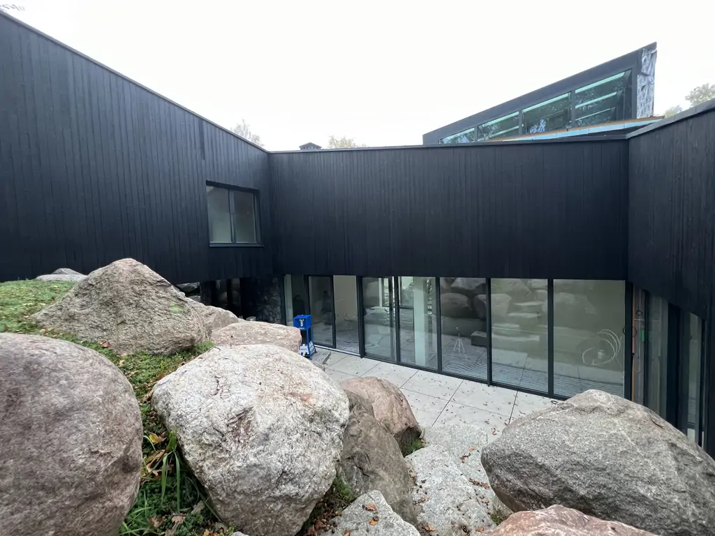 Boczna elewacja nowoczesnego domu wykończona czarnym drewnem, z kamiennym cokołem i głazami przy skarpie.