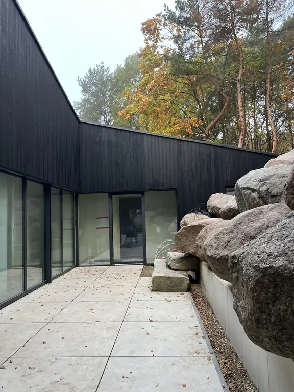 Dziedziniec nowoczesnego domu z czarną drewnianą elewacją, dużymi przeszkleniami i naturalnymi głazami wkomponowanymi w skarpę.