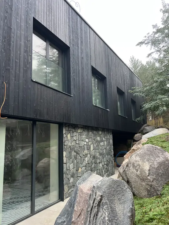 Narożna elewacja nowoczesnego domu z czarnym drewnem, kamieniem naturalnym i panoramicznymi przeszkleniami.