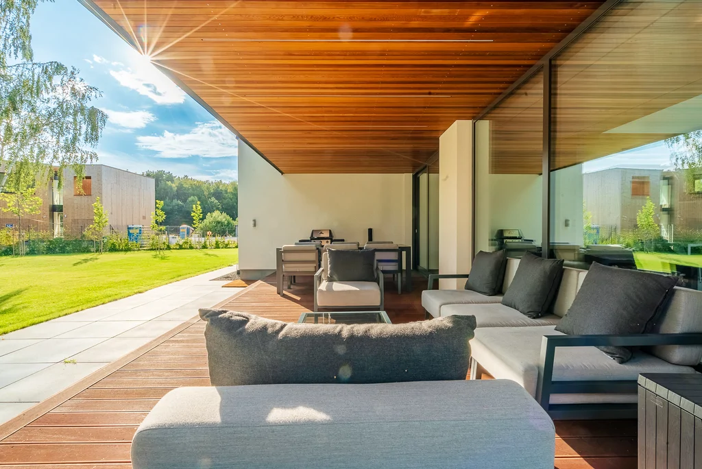 Zadaszony taras nowoczesnego domu z drewnianym sufitem, meblami wypoczynkowymi i widokiem na ogród.
