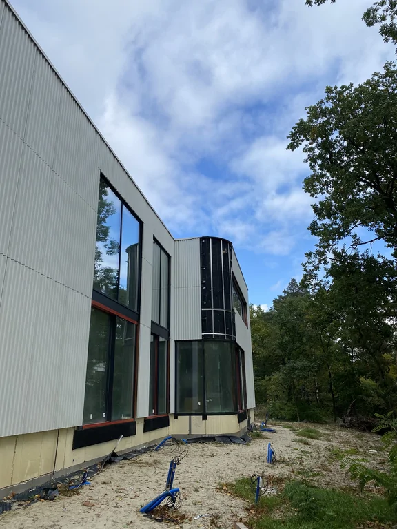 Boczna elewacja domu w trakcie realizacji z dużymi przeszkleniami, jasną okładziną elewacyjną i zaokrąglonym pionowym przeszkleniem