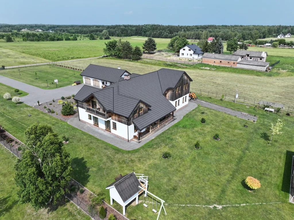 Widok z drona na posesję od strony ogrodu: dom, trawnik i budynek garażowy, na pierwszym planie huśtawka