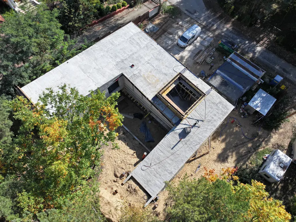 Widok z drona na żelbetowy budynek w stanie surowym z otwartym dziedzińcem, stropodachem i otoczeniem leśnym.