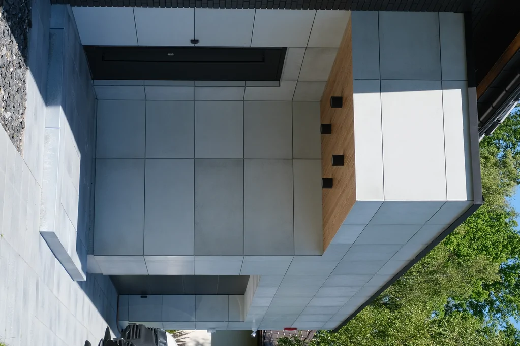 Detal strefy wejściowej: jasne płyty elewacyjne, podcień z drewnianą podbitką i oprawami oświetleniowymi.