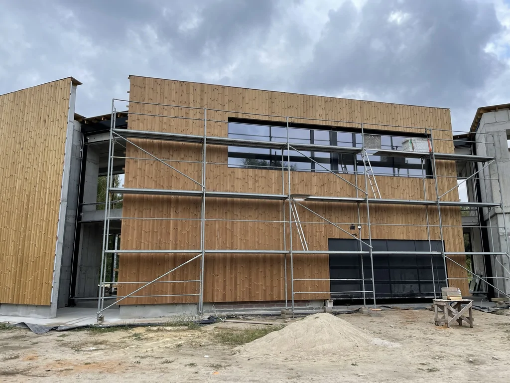 Zbliżenie na elewację: drewniana okładzina, długie przeszklenie i rusztowanie przy pracach wykończeniowych.