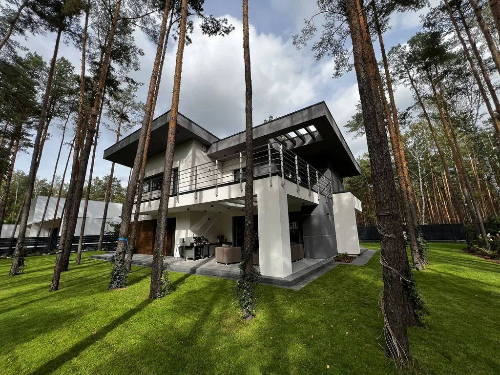 Widok od ogrodu: taras z meblami i grillem pod balkonem, nowoczesna bryła wśród sosen
