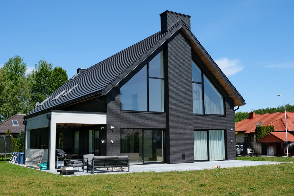 Widok na taras i elewację ogrodową: dom z ciemnej cegły, duże przeszklenia szczytowe i zadaszona strefa wypoczynku.