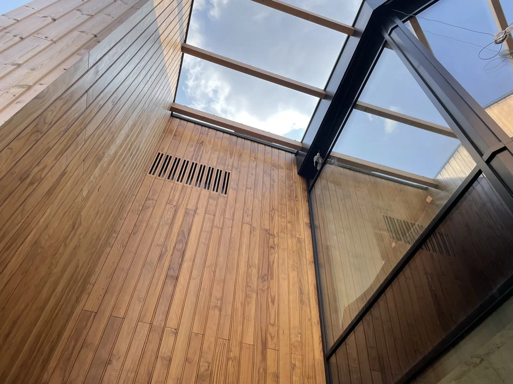 Detal atrium: drewniana ściana z kratką wentylacyjną oraz szklane zadaszenie na belkach konstrukcyjnych.