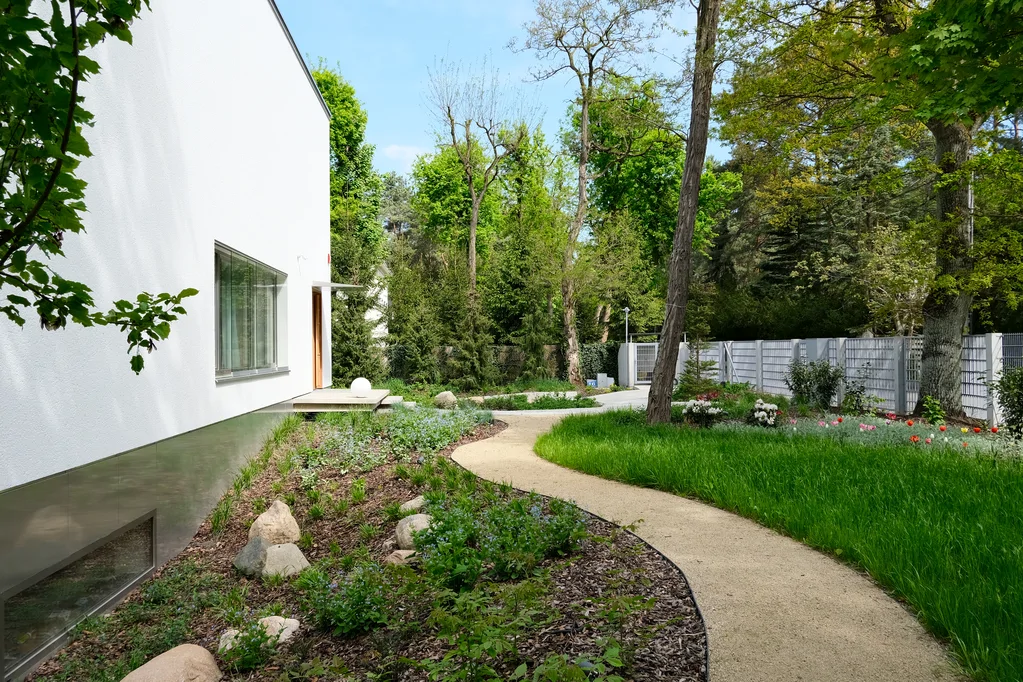 Widok na boczną ścieżkę ogrodową prowadzącą wzdłuż białej elewacji domu i zielonych rabat