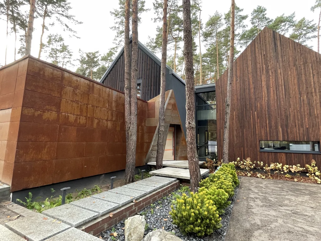 Strefa wejściowa nowoczesnego domu w Magdalence z elewacją cortenową i drewnianą bryłą w otoczeniu sosen