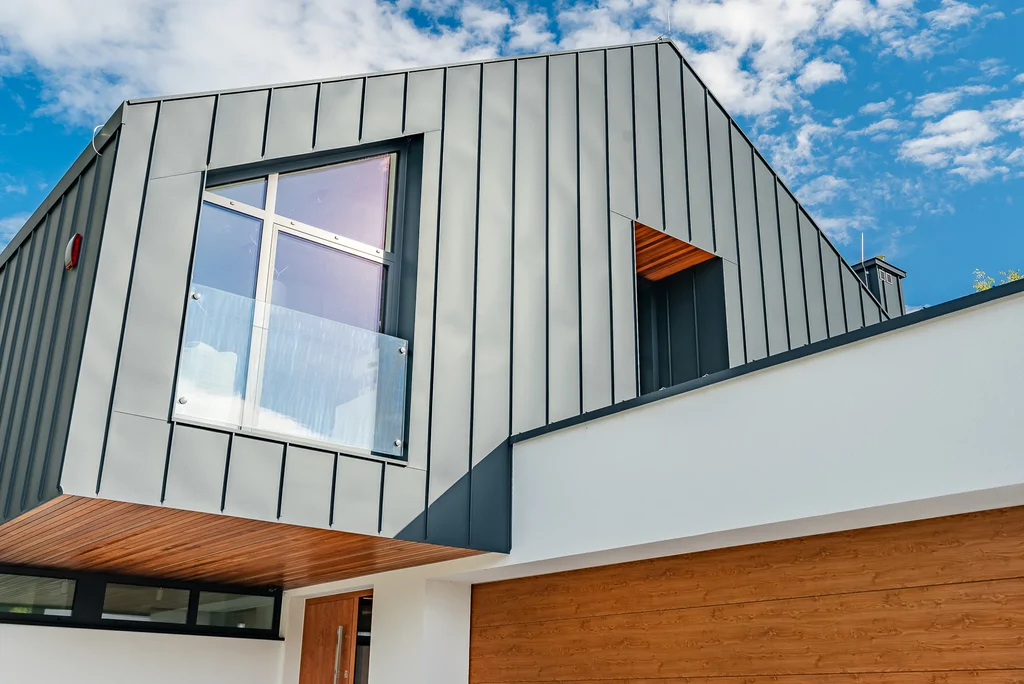 Zbliżenie górnej części elewacji z ciemną blachą na rąbek, oknem balkonowym i drewnianym podbiciem.