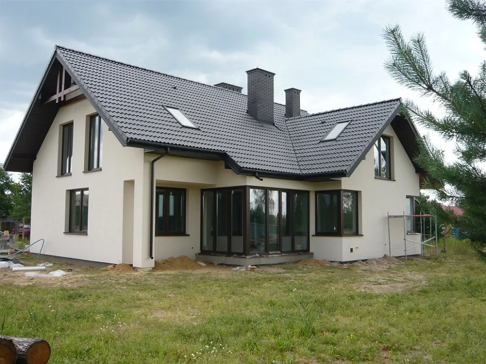 Dom jednorodzinny w stanie deweloperskim: jasna elewacja, ciemny dach i duże przeszklenia od strony ogrodu