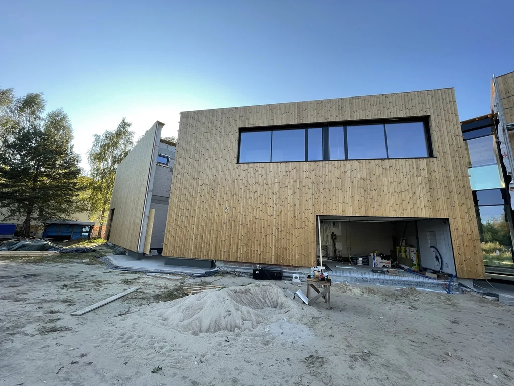 Boczna ściana w drewnie z poziomym oknem na piętrze oraz otworem garażowym; widoczne materiały i sprzęt budowlany.