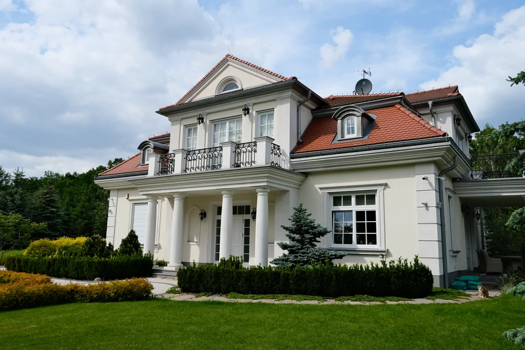 Dom jednorodzinny w stylu dworkowym, kolumny, biała elewacja, barierki, bonie na elewacji, dach z karpiówki, lukarna dachowa, ozdobne krzewy w strefie wejścia, 