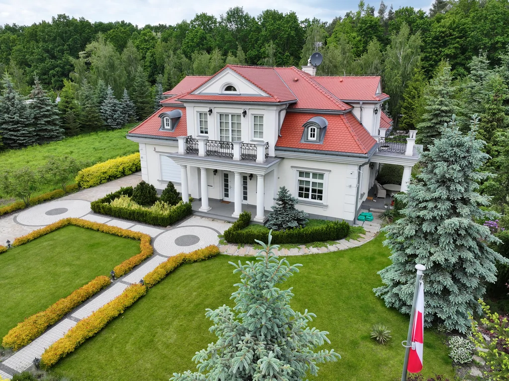 Widok z drona na dom jednorodzinny w stylu dworkowym z białą elewacją z boniami, kolumny okrągłe w strefie wejścia, na balkonie również kolumny i stalowa barierka, pokrycie dachu karpiówka, na dachu widoczne lukarny, przy domu kostka, działka otoczona drzewami 