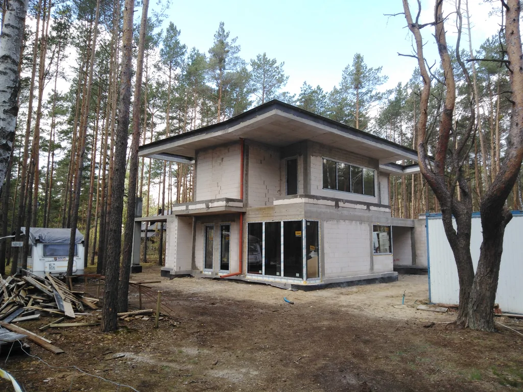 Front domu z zamontowanymi oknami i drzwiami tarasowymi, teren budowy wśród sosen