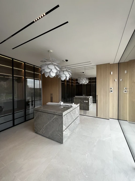 Centralna wyspa w kamieniu/spieku, zabudowa z ciemnymi witrynami i podświetleniem półek. Elegancki, “hotelowy” klimat.