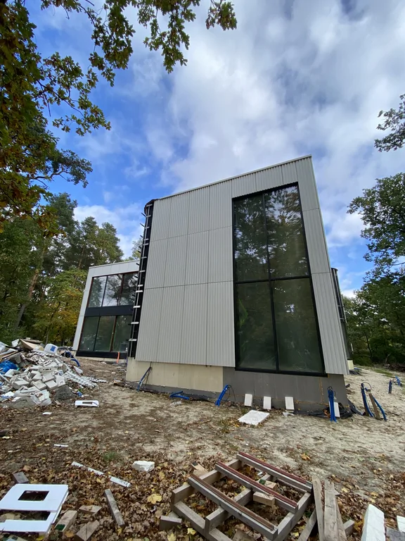 Narożnik nowoczesnego budynku z wysokim przeszkleniem na całą kondygnację, jasną ryflowaną elewacją i placem budowy wokół