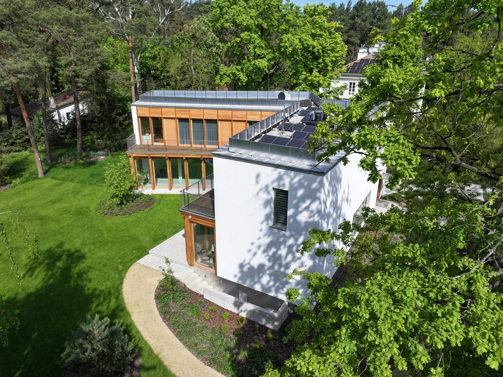 Ujęcie z drona na boczną część domu z białą bryłą, balkonem i panelami fotowoltaicznymi widocznymi na dachu