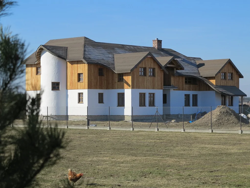 Konie na pastwisku przy ogrodzeniu; w tle duży dom w trakcie budowy z drewnianą elewacją