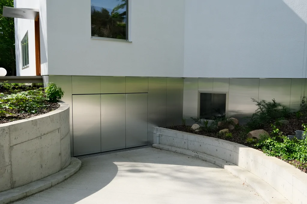 Ujęcie z poziomu podjazdu na bramę garażową w przyziemiu, stalowe okładziny i betonowe ściany oporowe