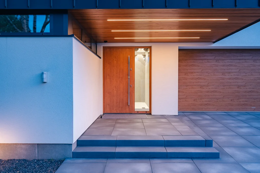 Wejście do domu o zmierzchu z podświetlaną drewnianą podsufitką, drewnianymi drzwiami i bramą garażową.