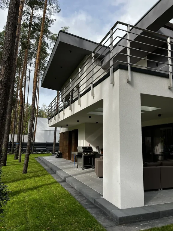 Taras przy domu: podcień, grill i strefa wypoczynku pod balkonem ze stalową balustradą