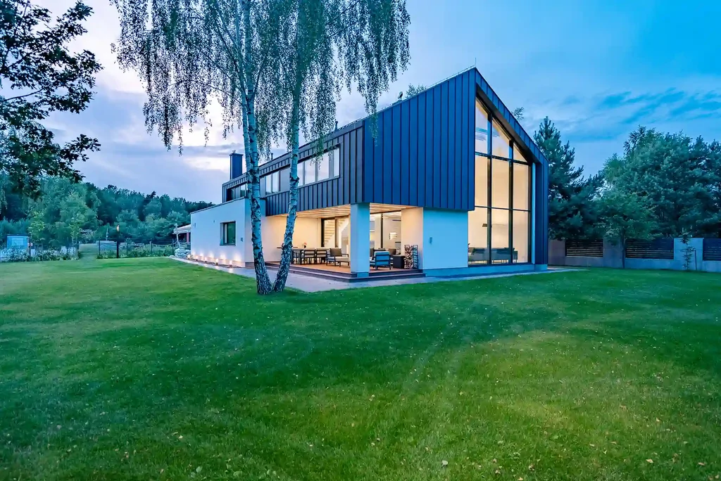 Wieczorne ujęcie nowoczesnego domu od strony ogrodu z wysokim przeszkleniem i oświetlonym tarasem.