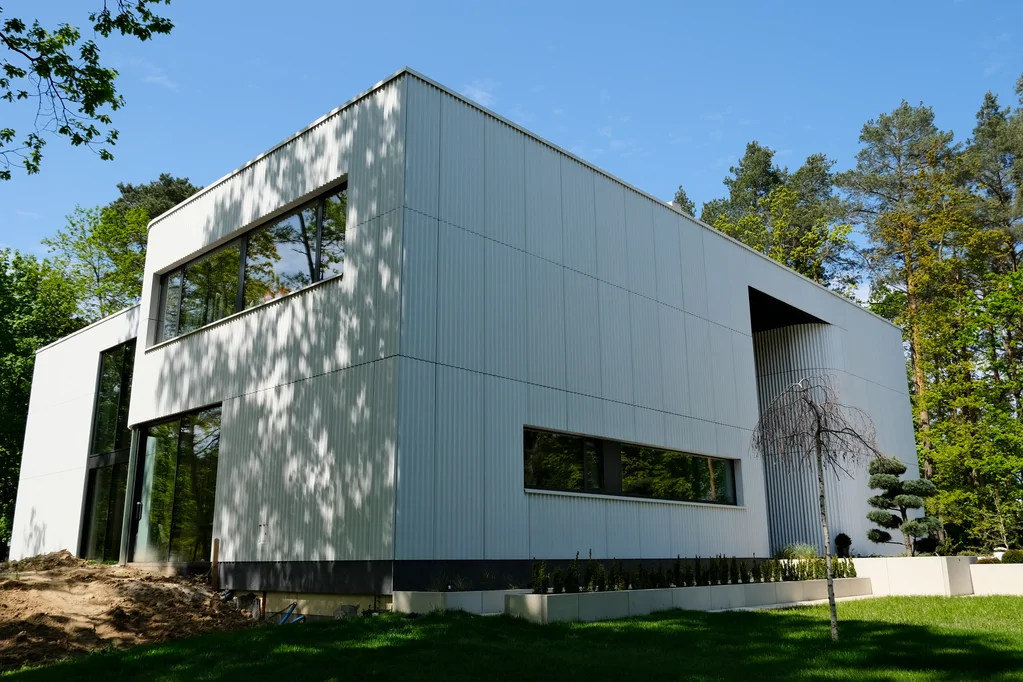 Ukośny widok na front i bok domu z jasną elewacją, poziomym oknem, cofniętym wejściem oraz zadbanym trawnikiem