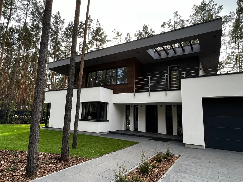 Front nowoczesnego domu w lesie: biała elewacja, drewno, duże okapy i podjazd przy garażu