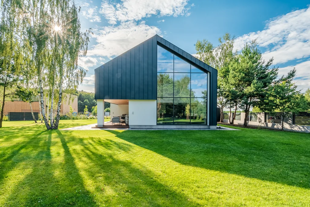 Szczytowa elewacja domu z wysokim przeszkleniem od podłogi do dachu i rozległym zielonym ogrodem.