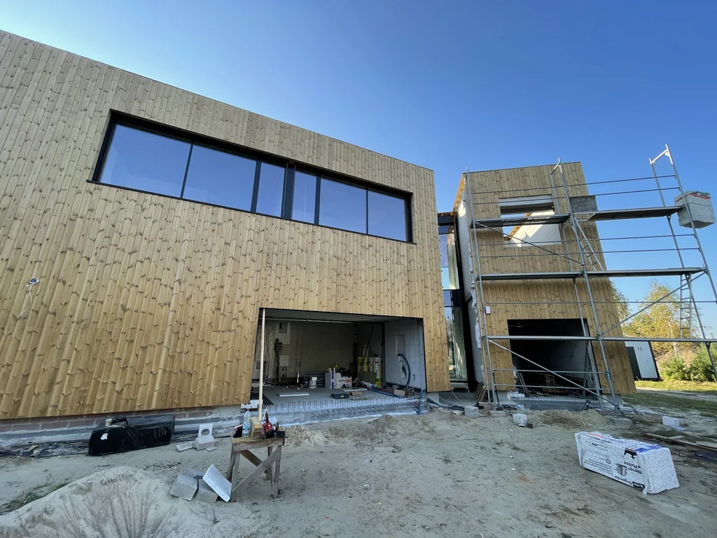 Elewacja drewniana z pasmowym oknem; otwarty garaż i prace montażowe na budowie.