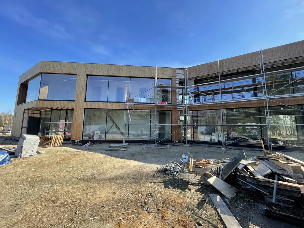 Panoramiczny widok fasady: drewniana okładzina, rozległe przeszklenia i rusztowanie przy pracach zewnętrznych.