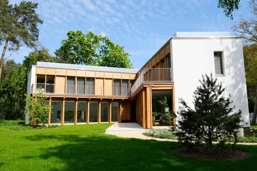 Ujęcie narożnika domu od strony ogrodu z białą bryłą, drewnianym podcieniem i sosną na pierwszym planie