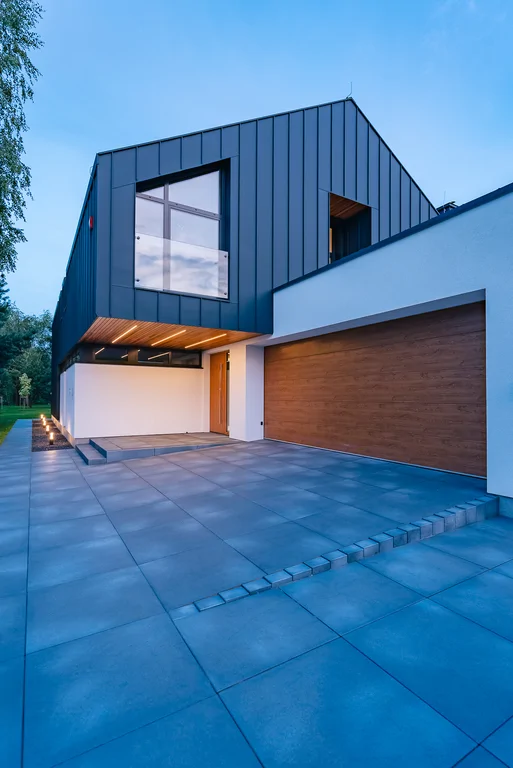 Frontowa elewacja domu o zmierzchu z szerokim podjazdem, garażem i podświetleniem pod nadwieszoną bryłą.