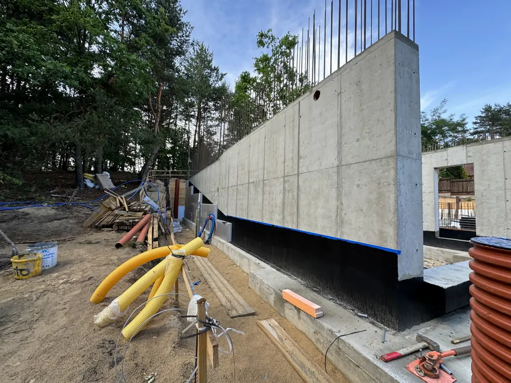 Długa żelbetowa ściana piwnicy z izolacją przeciwwodną i instalacjami prowadzonymi w gruncie.