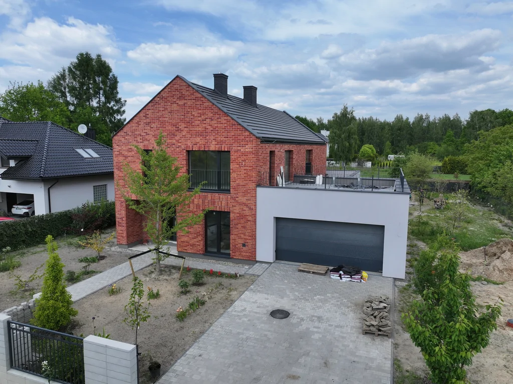 Centralny widok frontowy z drona na dom z ceglaną fasadą, garażem i młodą zielenią w ogrodzie frontowym