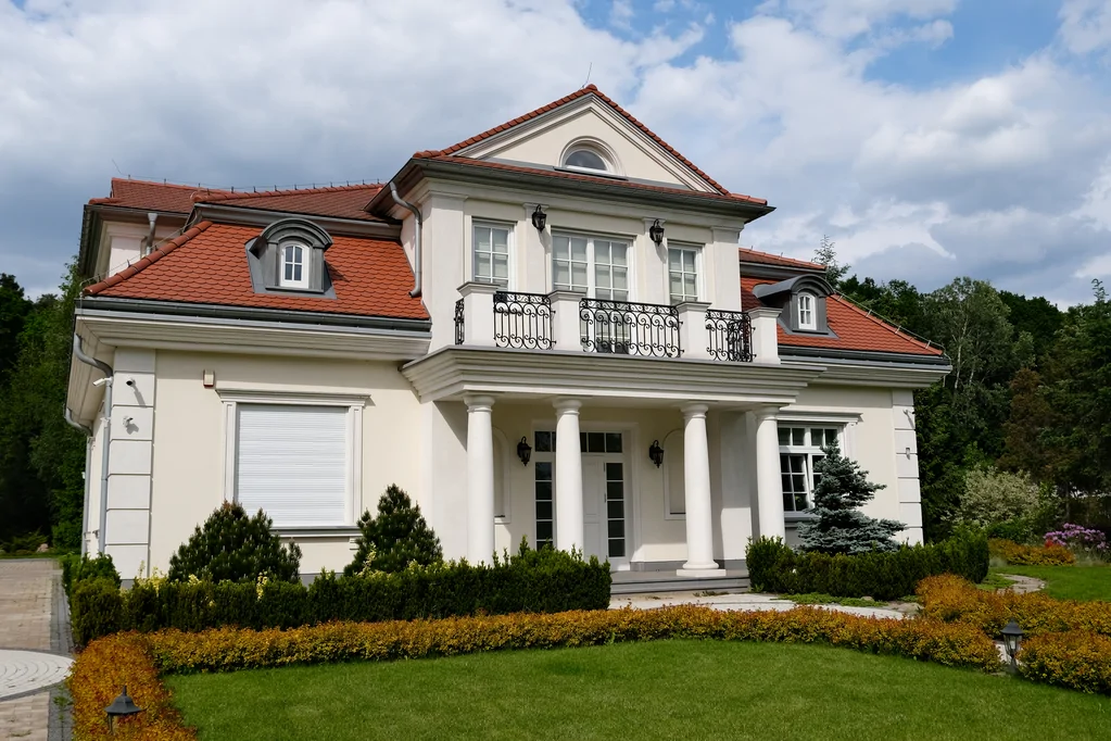 Dom jednorodzinny w stylu dworkowym, kolumny, biała elewacja, barierki na balkonie, bonie na elewacji, dach z karpiówki, lukarna dachowa, ozdobne krzewy w strefie wejścia, zasłonięte rolety