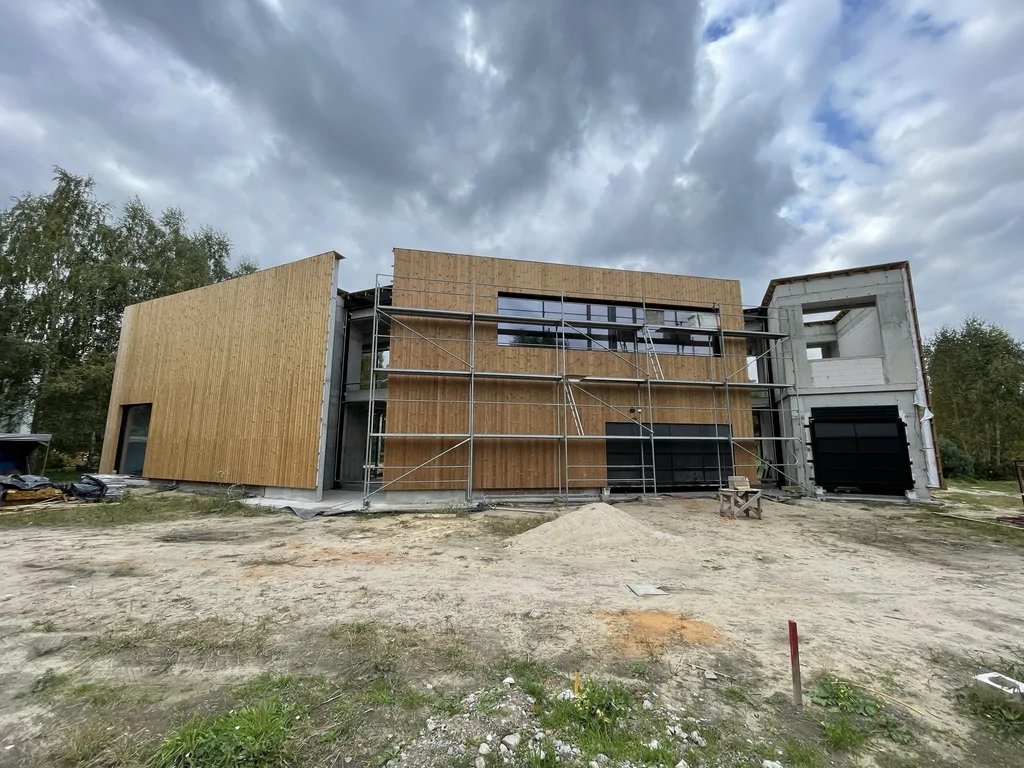 Front budynku z elewacją drewnianą i rusztowaniem – pasmo okien na piętrze oraz strefa garażu w trakcie montażu.