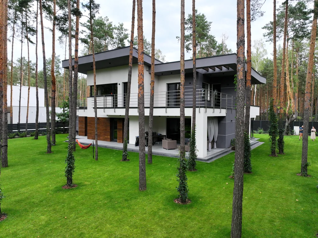 Ujęcie z góry: płaski dach domu z tarasem, bryła pośród wysokich sosen na ogrodzonej działce